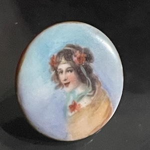 May include: A round, hand-painted porcelain brooch featuring a portrait of a woman with brown hair adorned with red flowers. The woman is wearing a yellow shawl against a light blue background. The brooch has a gold-colored rim.