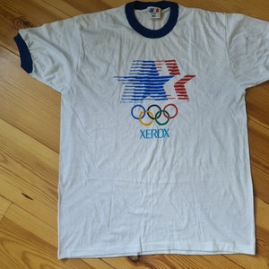 May include: White t-shirt with a blue and red star design, the Olympic rings, and the word "XEROX" printed on the front. The shirt has blue ringer sleeves.
