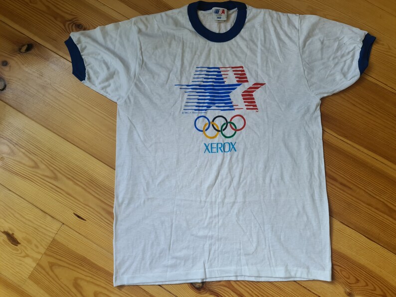 May include: White t-shirt with a blue and red star design, the Olympic rings, and the word "XEROX" printed on the front. The shirt has blue ringer sleeves.