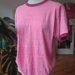 Puede incluir: Una camiseta rosa con cuello redondo y mangas cortas burdeos. La camiseta se exhibe en un maniquí. La tela tiene una textura jaspeada. El estilo general es informal y adecuado para el uso diario.