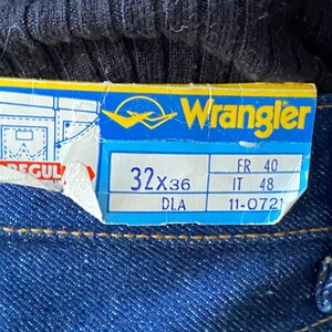 Pode incluir: Uma etiqueta de jeans Wrangler azul com texto branco. A etiqueta mostra o tamanho 32x36 e as medidas em tamanhos franc&ecirc;s, italiano e DLA. A etiqueta est&aacute; parcialmente rasgada.