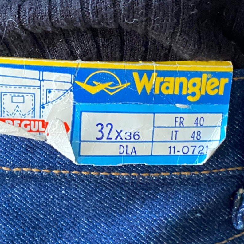 Pode incluir: Uma etiqueta de jeans Wrangler azul com texto branco. A etiqueta mostra o tamanho 32x36 e as medidas em tamanhos franc&ecirc;s, italiano e DLA. A etiqueta est&aacute; parcialmente rasgada.