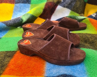 Puede incluir: Un par de zapatillas de pana marrón con un logotipo en forma de corazón en el lateral. Las zapatillas están sobre una manta colorida.