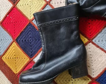 May include: A pair of black leather ankle boots with a low block heel. The boots have a decorative stitching detail around the top of the shaft. The boots are sitting on a colorful crocheted blanket.