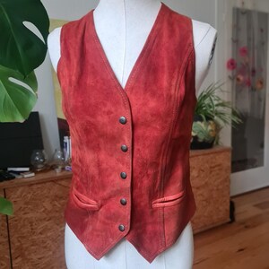 May include: A vintage red suede vest with a V-neck and a button-up front. The vest has two front pockets and is displayed on a mannequin. The vest is a reddish-orange color and appears to be in good condition.