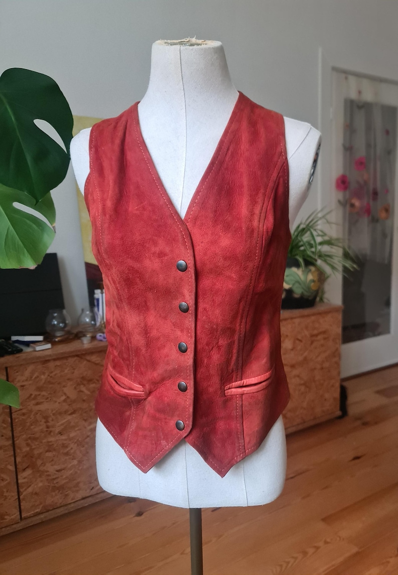 May include: A vintage red suede vest with a V-neck and a button-up front. The vest has two front pockets and is displayed on a mannequin. The vest is a reddish-orange color and appears to be in good condition.
