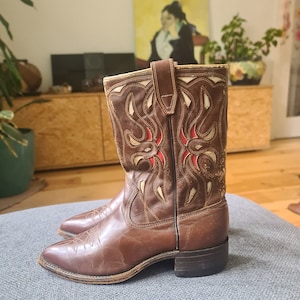 May include: Brown leather cowboy boots with intricate white and red stitching on the shaft. The boots have a pointed toe and a low heel.