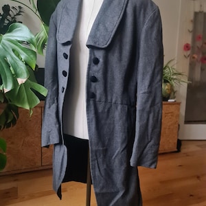May include: A grey wool coat with a wide collar and three black buttons. The coat is displayed on a mannequin. The coat has a long, tailored design, with a slight flare at the bottom. The coat is a classic style.