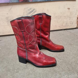 Könnte beinhalten: Ein Paar rote Leder-Cowboystiefel mit schwarzen Sohlen und Absätzen. Die Stiefel haben dekorative Nähte und Zuglaschen. Die Stiefel sind mittelhoch und haben eine quadratische Spitze.