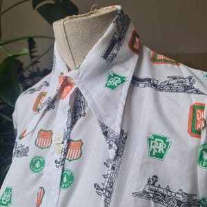 May include: White collared shirt featuring a repeating pattern of black train illustrations and orange and green railroad logos. The shirt has short sleeves and a button-down front. The collar is white with a black train illustration.
