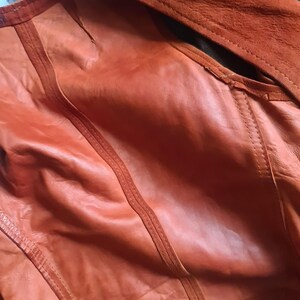 May include: Close-up of an orange leather jacket with a suede collar. The jacket features detailed stitching and a textured surface. The color is a warm, reddish-orange, and the garment appears to be well-crafted.