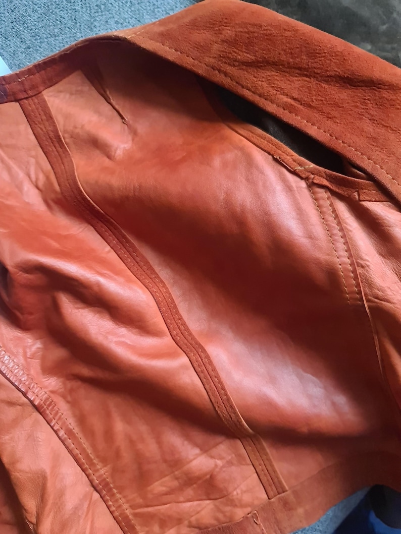 May include: Close-up of an orange leather jacket with a suede collar. The jacket features detailed stitching and a textured surface. The color is a warm, reddish-orange, and the garment appears to be well-crafted.