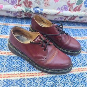 May include: A pair of burgundy leather Oxford shoes with black laces. The shoes have a classic design with a yellow stitched sole. The shoes are placed on a blue and white patterned fabric.