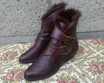 May include: A pair of brown leather ankle boots with fur trim. The boots have a buckle closure and a low heel. The boots are displayed on a floral patterned surface against a grey brick wall.