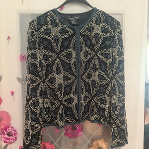 Könnte beinhalten: Schwarze Jacke mit aufwendigem Blumenmuster aus weißen und blauen Perlen. Die Jacke hat einen runden Ausschnitt und eine offene Front. Das florale Design ist symmetrisch und wiederholt sich über den Stoff. Die Jacke hängt an einem schwarzen Kleiderbügel.