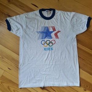 May include: White t-shirt with a blue and red star design, the Olympic rings, and the word "XEROX" printed on the front. The shirt has a blue ringer.