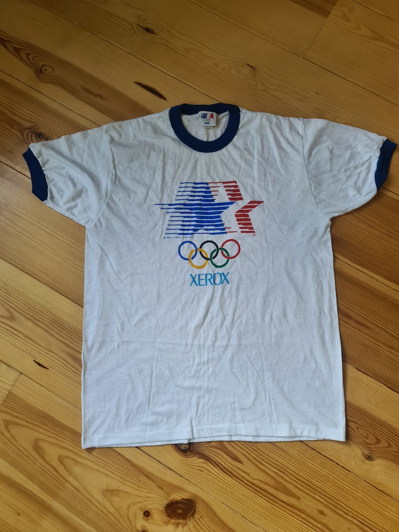 May include: White t-shirt with a blue and red star design, the Olympic rings, and the word "XEROX" printed on the front. The shirt has a blue ringer.