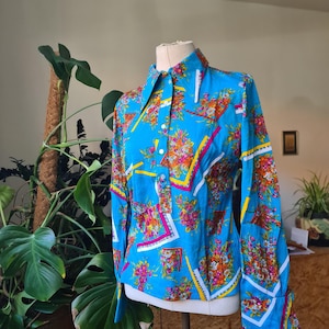 Könnte beinhalten: Ein Vintage-Langarmhemd mit einem leuchtenden Blumenmuster in Rosa, Orange und Gelb auf hellblauem Hintergrund. Das Hemd hat einen spitzen Kragen und geknöpfte Manschetten, präsentiert auf einer Schaufensterpuppe.