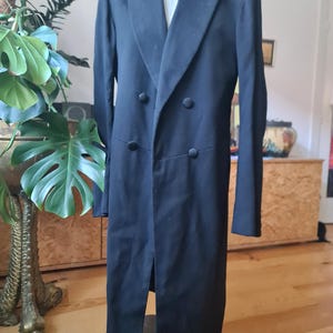 May include: A black double-breasted tailcoat on a mannequin. The coat has a notched lapel, two rows of buttons, and a long, split back. The dark fabric garment is suitable for formal events. The coat's length is approximately 120 cm.