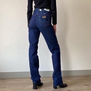 Pode incluir: Um par de cal&ccedil;as de ganga azul escuro com cintura alta e perna direita. As cal&ccedil;as t&ecirc;m um design cl&aacute;ssico de cinco bolsos e um remendo de couro na parte de tr&aacute;s da cintura com o logotipo da Wrangler.