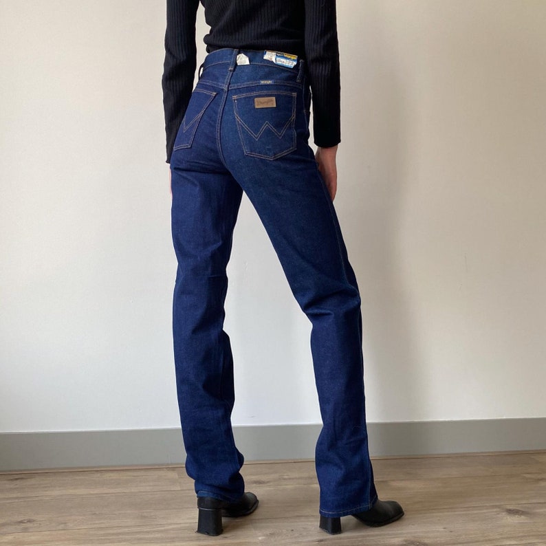 Pode incluir: Um par de cal&ccedil;as de ganga azul escuro com cintura alta e perna direita. As cal&ccedil;as t&ecirc;m um design cl&aacute;ssico de cinco bolsos e um remendo de couro na parte de tr&aacute;s da cintura com o logotipo da Wrangler.