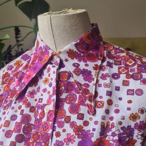 Könnte beinhalten: Ein Vintage-Hemd mit einem lebendigen Blumenmuster in Pink-, Lila- und Orangetönen auf weißem Grund. Das Hemd hat einen klassischen Kragen und einen einzelnen sichtbaren Knopf. Das florale Design umfasst geometrische Formen.