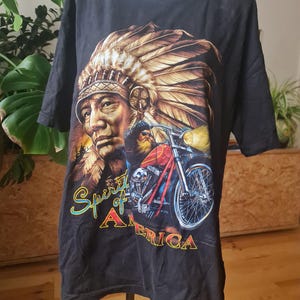Pode incluir: Camiseta preta com um desenho de um chefe nativo americano com um cocar de penas e uma motocicleta vermelha. Inclui o texto "Spirit of America". A camiseta é exibida em um manequim.