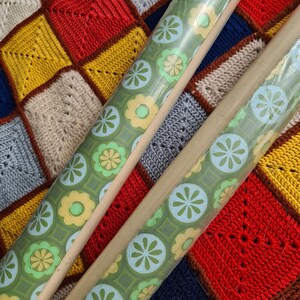 Puede incluir: Dos rollos de papel de regalo verde con estampado floral con detalles en azul claro y amarillo. Los rollos descansan sobre una colorida manta de ganchillo patchwork con cuadrados rojos, amarillos, azules y beige.