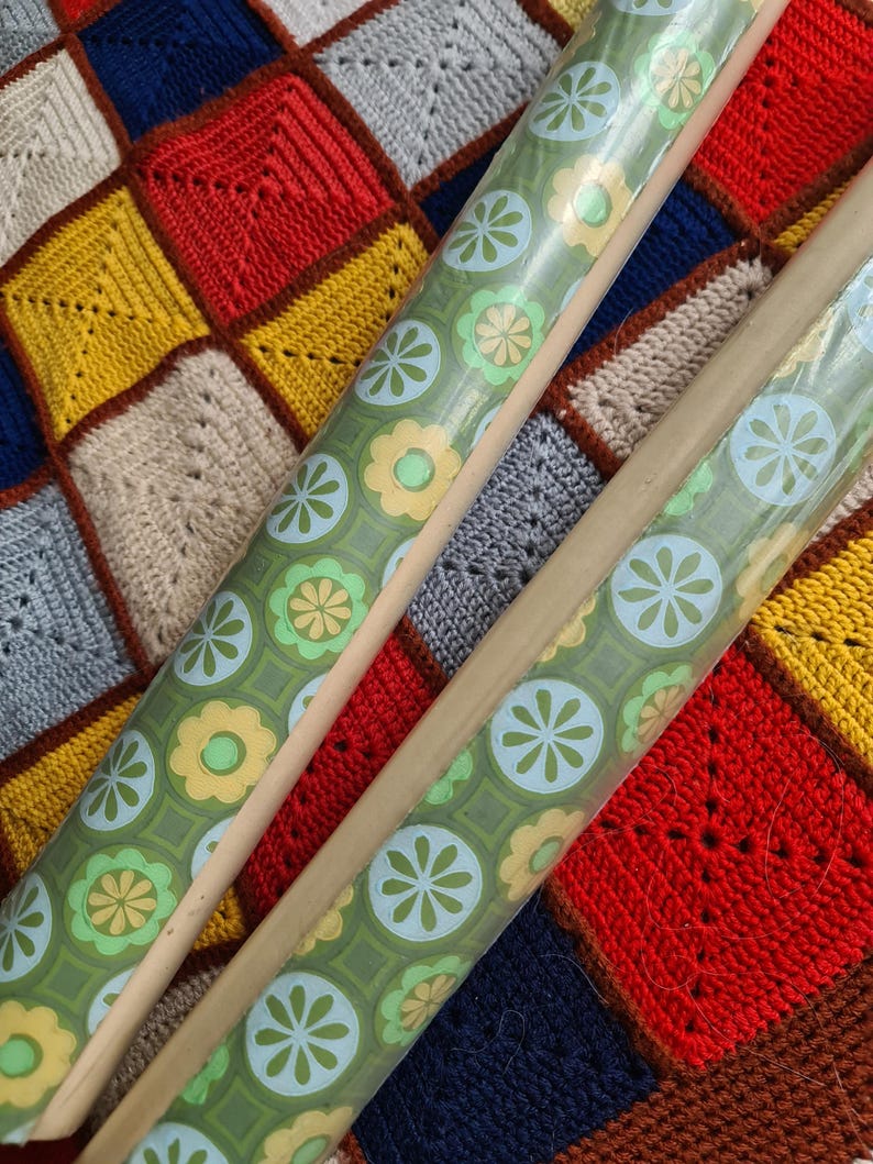 Puede incluir: Dos rollos de papel de regalo verde con estampado floral con detalles en azul claro y amarillo. Los rollos descansan sobre una colorida manta de ganchillo patchwork con cuadrados rojos, amarillos, azules y beige.