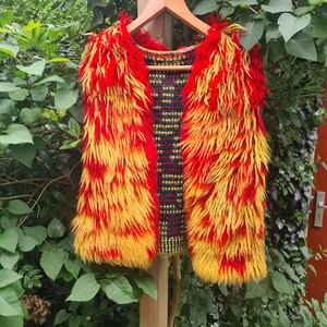 Puede incluir: Un chaleco de piel sintética rojo y amarillo con un forro estampado en negro y dorado. El chaleco está colgado en una percha de madera.