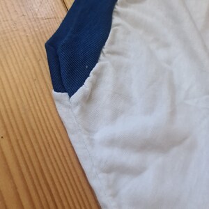 May include: Close-up of a white t-shirt with a blue and white color block design. The shirt has a ribbed collar and a blue trim around the neck.