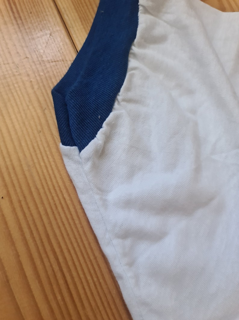 May include: Close-up of a white t-shirt with a blue and white color block design. The shirt has a ribbed collar and a blue trim around the neck.