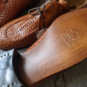 May include: Close-up of the sole of a brown leather shoe with a woven pattern. The sole is stamped with the text "GENUINE LEATHER" in a circle.
