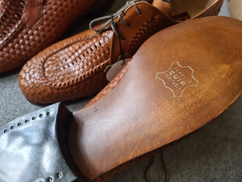 May include: Close-up of the sole of a brown leather shoe with a woven pattern. The sole is stamped with the text "GENUINE LEATHER" in a circle.