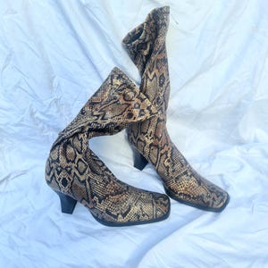 May include: A pair of knee-high boots with a snakeskin pattern in shades of brown, tan, and black. The boots have a square toe and a small, black heel. The boots are made of a textured material.