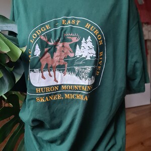 May include: Green t-shirt with a circular graphic featuring a moose, trees, and the text "East Huron River" and "Huron Mountains, Skanee, Michigan." The shirt is made of a soft material.