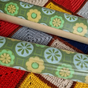 Puede incluir: Dos rollos de papel pintado vintage con un patr&oacute;n floral repetido en tonos verdes, amarillos y azules. El papel pintado est&aacute; enrollado en tubos de cart&oacute;n. El fondo es un colorido patchwork de cuadrados de ganchillo.