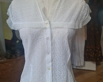 May include: White, short-sleeved blouse with a floral lace pattern. The blouse features a pleated collar and cap sleeves. It is buttoned down the front and displayed on a mannequin.