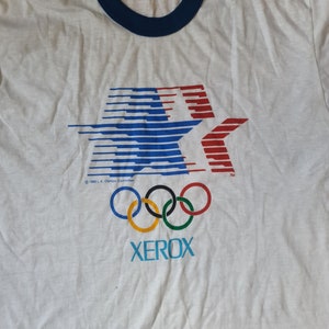 May include: White t-shirt with a blue and red star graphic, the Olympic rings, and the word "XEROX" printed on it. The shirt has a blue ringer neck and a tag that says "1984 L.A. Olympic Committee".