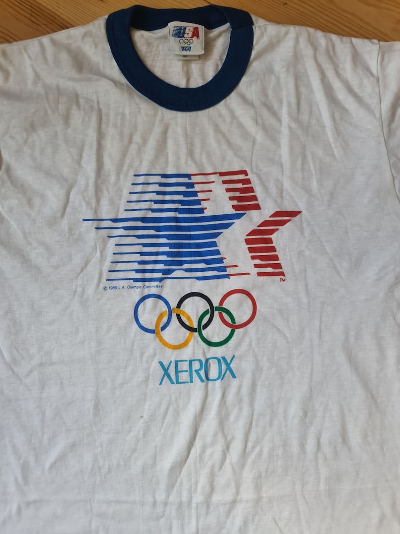 May include: White t-shirt with a blue and red star graphic, the Olympic rings, and the word "XEROX" printed on it. The shirt has a blue ringer neck and a tag that says "1984 L.A. Olympic Committee".