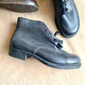 Puede incluir: Botines de cuero negro con cierre de cordones y aspecto texturizado. Las botas tienen una suela resistente y se muestran sobre una superficie marrón claro. Se ven zapatos de cuero marrón adicionales en el fondo.