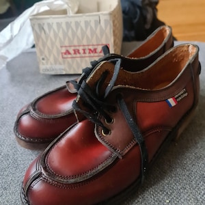 May include: A pair of brown leather lace-up shoes with a red and white stripe on the side. The shoes have a black lace and a brown leather sole. The shoes are labelled "Arima" and "Uniquely The College".