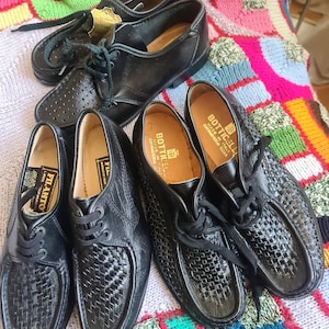 Pode incluir: Quatro pares de sapatos de couro preto com detalhes em tecido. Os sapatos têm cadarços e estão sobre um cobertor de malha.