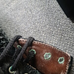 May include: Close-up of a black leather boot with brown leather lining. The boot has a lace-up closure and metal eyelets. The lining has several small, circular holes with a turquoise-colored border.