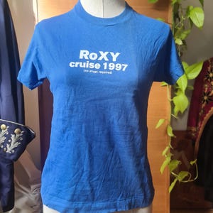 Könnte beinhalten: Ein blaues T-Shirt mit dem Text "ROXY cruise 1997 (no drugs required)" in Weiß. Das T-Shirt wird auf einer Schaufensterpuppe präsentiert. Das T-Shirt hat kurze Ärmel und einen Rundhalsausschnitt.