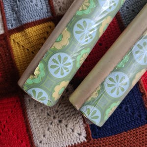 Puede incluir: Dos rollos de papel pintado vintage con un estampado floral verde, azul y amarillo. El papel pintado est&aacute; envuelto en pl&aacute;stico transparente y tiene un reverso marr&oacute;n claro. Los rollos descansan sobre una colorida manta de ganchillo.