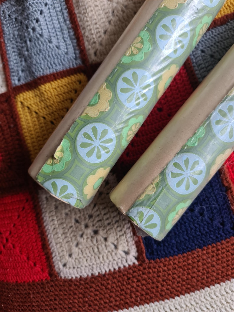 Puede incluir: Dos rollos de papel pintado vintage con un estampado floral verde, azul y amarillo. El papel pintado est&aacute; envuelto en pl&aacute;stico transparente y tiene un reverso marr&oacute;n claro. Los rollos descansan sobre una colorida manta de ganchillo.
