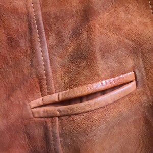 May include: Close-up of a brown suede jacket, showcasing the texture and stitching details. The image highlights a pocket with a leather trim. The jacket's color is a warm, earthy tone.