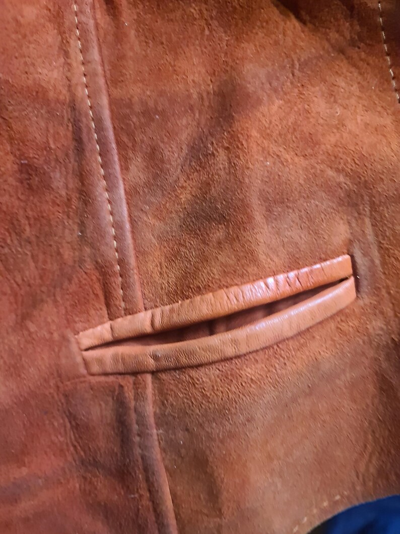 May include: Close-up of a brown suede jacket, showcasing the texture and stitching details. The image highlights a pocket with a leather trim. The jacket's color is a warm, earthy tone.