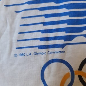 May include: White t-shirt with a blue and white striped design. The shirt has the text "&copy; 1980 L.A. Olympic Committee" and the Olympic rings in blue, yellow, black, and green.